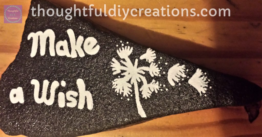 Black Spray Painted triangle shape stone with final layer of 'Make a Wish' Sentiment with white Paint Marker and finer details of the dandelion done with white paint pen.