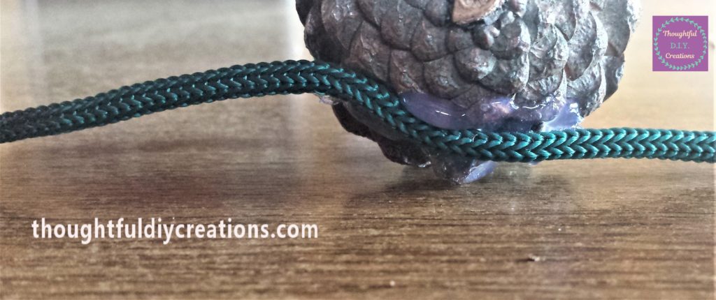 Hot glue on the back of the pine cone and attached to the string.