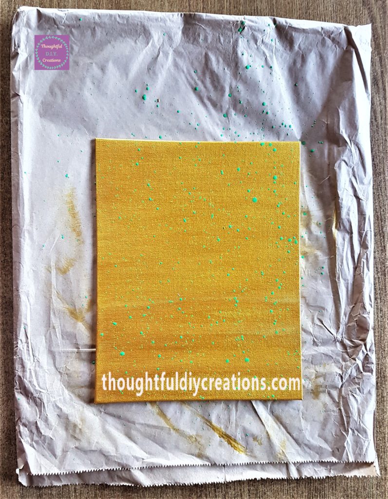 Brown paper bag on the table to protect it, canvas on top of the bag. Green Splashes added to the canvas by tapping a paintbrush over it.