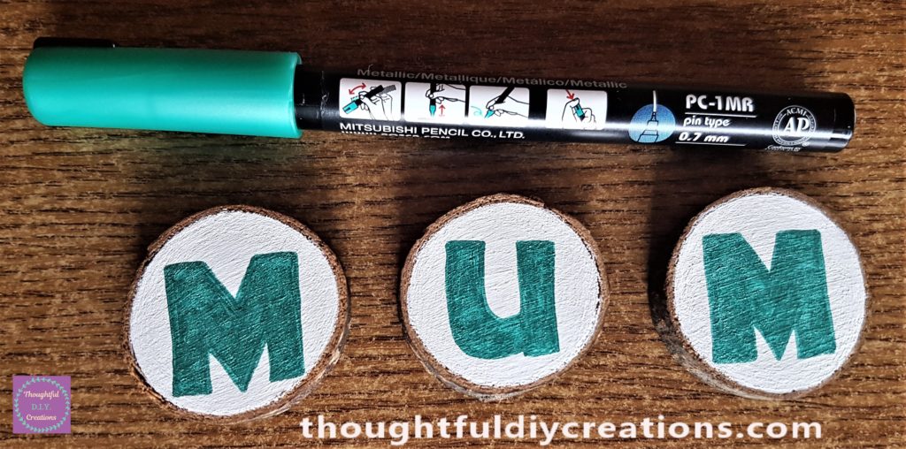 The word 'MUM' written and coloured in with the green paint pen on the wood slices.