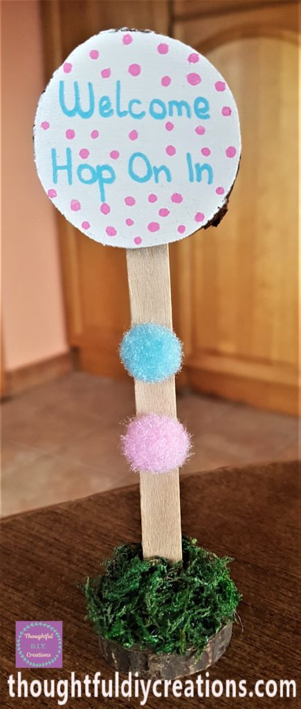 The painted wood slice hot glued to the top of the lollipop stick. A blue and pink pom pom hot glued to the lollipop stick.