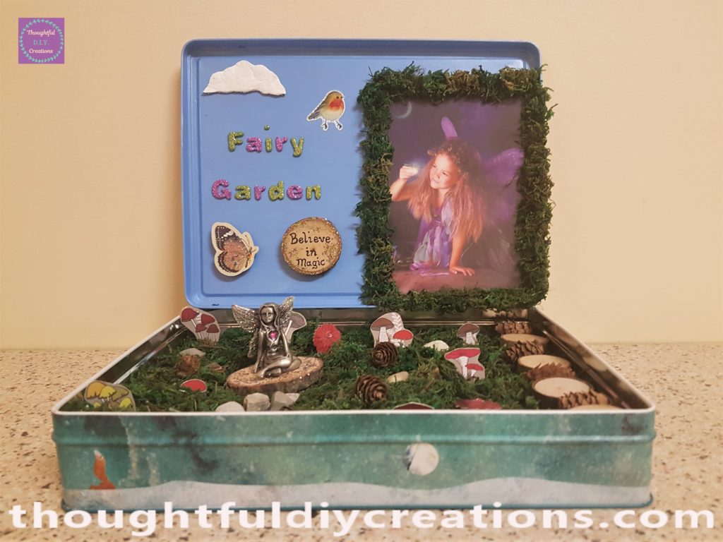 A robin sticker added to the sky on the fairy garden lid. Hot glue put along the base of the lid and attached to the back edge of the biscuit tin. More hot glue added at the back.